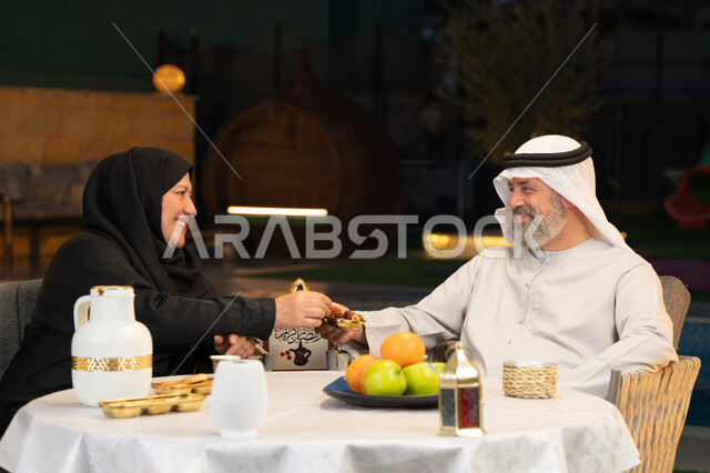 زينة وديكور المناسبات والأعياد الاسلامية، أجواء عائلية سعيدة في شهر رمضان المبارك، زوجان عربيان خليجيان اماراتيان يقومان بتناول التمربإيماءات السعادة والسرور، قضاء وقت ممتع في جلسة خارجية في احدى الشاليهات
