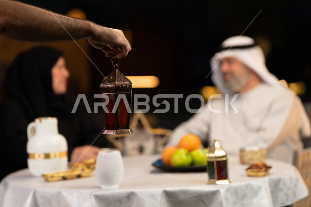 ديكورات وزينة المناسبات والأعياد الاسلامية، تبادل الأحاديث والتسامر بين الأفراد، صورة مقربة ليد رجل يحمل فانوس معدني مضيء، زوجان عربيان خليجيان اماراتيان يقضيان أوقات ممتعة في جلسة خارجية في احدى الشاليهات، أجواء عائلية سعيدة في شهر رمضان المبارك
