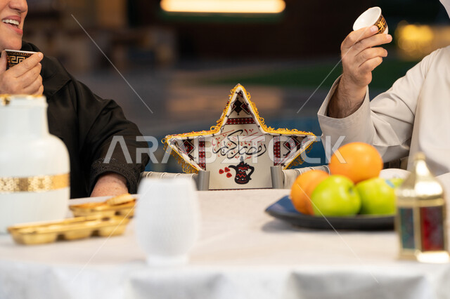 إيماءات السرور والسعادة، زينة وديكورات المناسبات والأعياد الاسلامية، أجواء عائلية سعيدة في شهر رمضان المبارك، صورة مقربة لأيدي زوجين عربيين خليجيين إماراتيين يتناولان القهوة العربية، جلسة خارجية في احدى الشاليهات الراقية