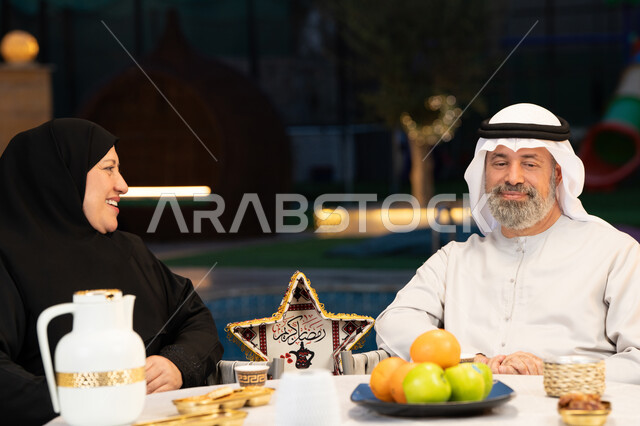 قضاء أوقات ممتعة في الهواء الطلق، ديكورات وزينة المناسبات والأعياد الاسلامية، أجواء عائلية سعيدة في شهر رمضان المبارك، تبادل الأحاديث والتسامر بين الزوجين، أسرة عربية خليجية إماراتية في جلسة خارجية في احدى الشاليهات الراقية