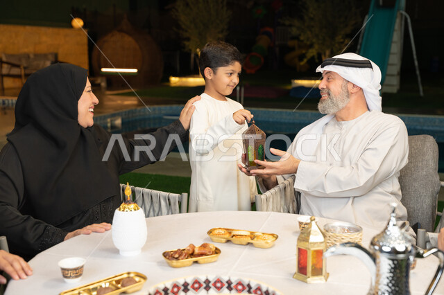إيماءات السعادة والسرور بالاجتماع العائلي، الحلويات الرمضانيه المتنوعه والشهية، زينة وديكور المناسبات والأعياد الاسلامية، أسرة عربية خليجية إماراتية تقضي وقت ممتع في جلسة خارجية في احدى الشاليهات، ولد يحمل بيده فانوس معدني مضيء