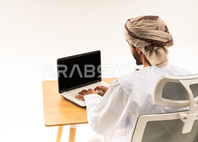 مفهوم إدارة العمل عن بعد، الإشراف على سير الأعمال عبر جهاز الحاسب المحمول، الاستفادة من التقنيات الحديثة في القطاع المكتبي والإداري، بورتريه من الخلف لرجل عربي خليجي عماني يرتدي الدشداشه والعمامه يجلس على طاولة المكتب ينظر بإيماءات الاندماج، خلفية بيضاء