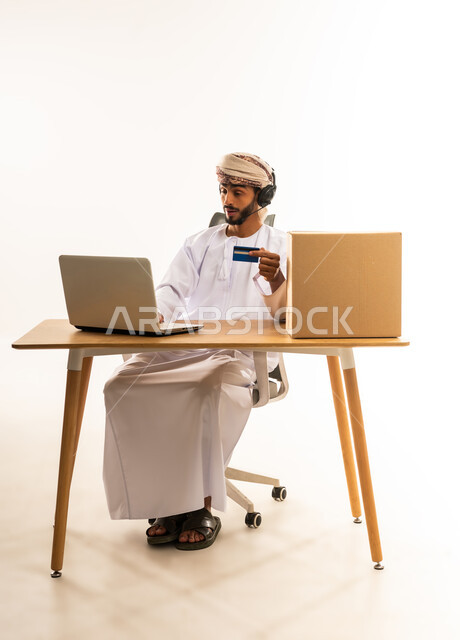 Easy and convenient remote shopping, electronic payment process using credit card, purchasing through applications and websites, the concept of online shopping, portrait of an Arab Gulf Omani man wearing traditional thobe and turban sitting behind a desk in front of a laptop, white background