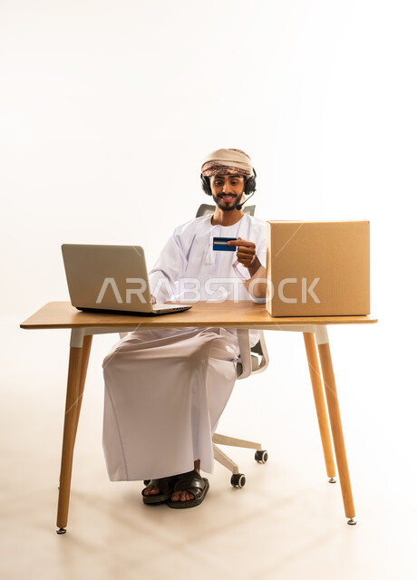 Electronic payment process using credit card, purchasing through applications and websites, the concept of online shopping, easy and convenient remote shopping, portrait of an Arab Gulf Omani man wearing traditional thobe and turban sitting behind a desk in front of a laptop, white background