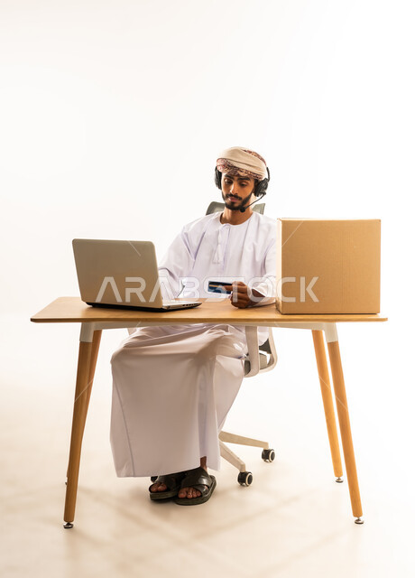 الشراء عبر تطبيقات ومواقع الانترنت، عملية الدفع الالكتروني بواسطة بطاقة الائتمان، مفهوم التسوق أونلاين، التبضع عن بعد بسهولة ويسر، بورتريه لرجل عربي خليجي  عماني يرتدي الثوب التقليدي و العمامة يجلس خلف المكتب أمامه الحاسوب المحمول، خلفية بيضاء