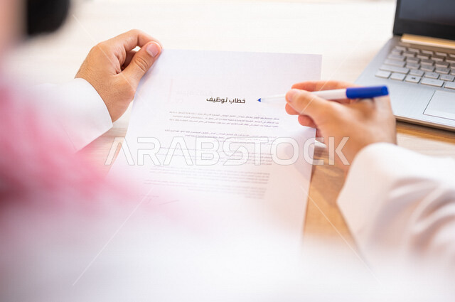 A Saudi Gulf Arab man sitting on a laptop and holding a hand an employment letter, reading an employment letter, a secretary managing paperwork, the concept of employment and work, office work