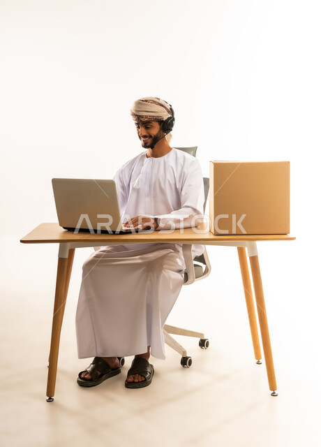 مهن ووظائف مكتبيه عمانية، مفهوم ادارة الاعمال عن بعد، بورتريه لشاب عربي خليجي عماني يرتدي الدشداشة والعمامة و السماعات يجلس خلف طاولة خشبية وامامه حاسوب، استخدام الأجهزة الحديثة والتقنيات المتطورة، خلفية بيضاء