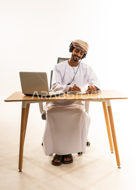 The concept of diligence and completion of required tasks, integrating modern and technological techniques into the administrative and office field, the concept of remote business management, a portrait of a young Arab Gulf Omani man wearing a dishdasha, turban and headphones, sitting behind a wooden table with a computer in front of him taking notes, white background