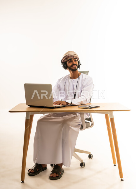 ايماءات السعاده و السرور، دمج التقنيات الحديثة والتكنولوجية بالمجال الإداري والمكتبي، مفهوم ادارة الاعمال عن بعد، ، بورتريه لشاب عربي خليجي عماني يرتدي الدشداشة والعمامة و السماعات يجلس خلف طاولة خشبية وامامه حاسوب يقوم بالنظر الى الاعلى، خلفية بيضاء