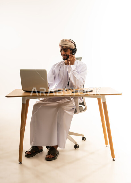 دمج التقنيات الحديثة والتكنولوجية بالمجال الإداري والمكتبي، مفهوم ادارة الاعمال عن بعد، ، بورتريه لشاب عربي خليجي عماني يرتدي الدشداشة والعمامة و السماعات يجلس خلف طاولة خشبية وامامه حاسوب يقوم باجراء مكالمة هاتفية، خلفية بيضاء