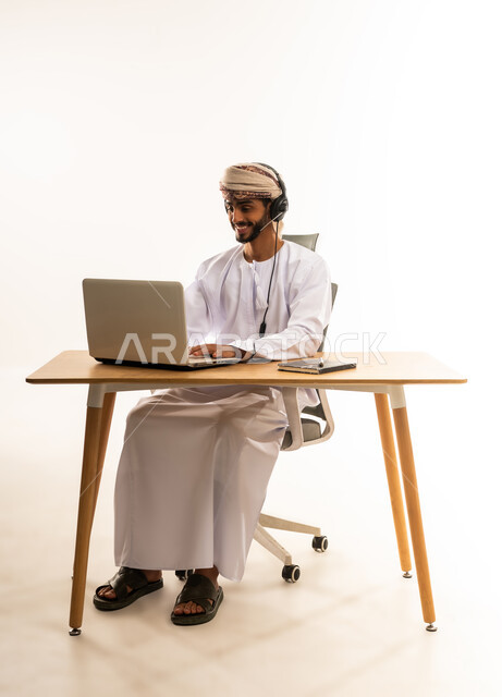 مفهوم ادارة الاعمال عن بعد، مهن ووظائف مكتبيه عمانية، بورتريه لشاب عربي خليجي عماني يرتدي الدشداشة والعمامة و السماعات يجلس خلف طاولة خشبية وامامه حاسوب، استخدام الأجهزة الحديثة والتقنيات المتطورة، خلفية بيضاء