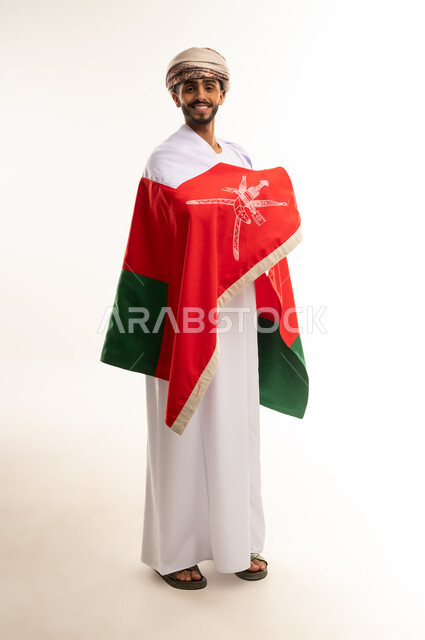 النظر الى الكاميرا بإيماءات السرور و الفرح, الاعتزاز بالانتماء للهوية، الاحتفال بذكرى اليوم الوطني العماني، الفخر بالرموز والراية العمانية، بورتريه لشاب عربي خليجي عماني يرتدي  العمامة والدشداشة  يضع العلم على اكتافه، الجسم بالطول الكامل، خلفية بيضاء