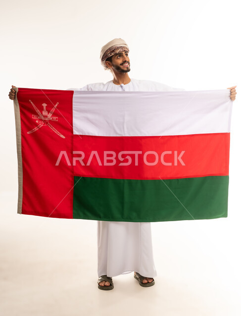 المشاركة بالاعياد والمناسبات، الفخر والاعتزاز بالهوية الوطنية ، العلم الوطني للبلاد، بورتريه لرجل عربي خليجي عماني مبتسم يرتدي الدشداشة و العمامه يقوم بامساك العلم و ينظر الى شيء ما  ،الجسم بالطول الكامل، خلفية بيضاء