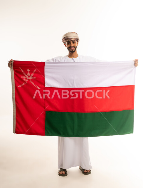 ثقافات و رموز وطنية، الفخر والاعتزاز بالهوية الوطنية ، العلم الوطني للبلاد، بورتريه لرجل عربي خليجي عماني مبتسم يرتدي الدشداشة و العمامه يحتفل بذكرى اليوم الوطني ،الجسم بالطول الكامل، خلفية بيضاء