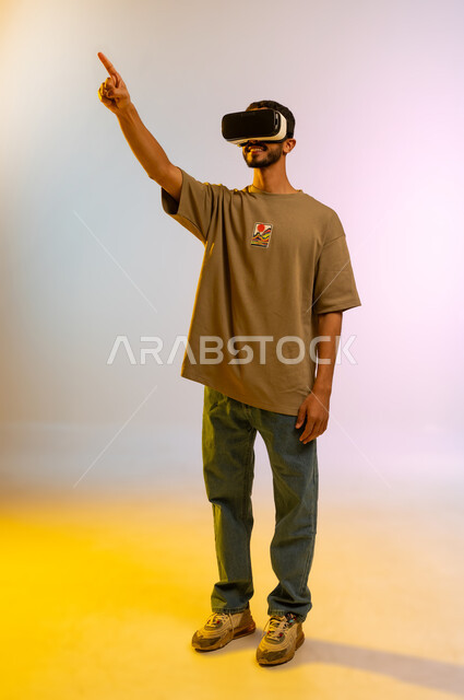 التفاعل ومحاكاة العالم الخيالي، رجل عماني يقوم بمشاهدة أفلام عبر نظارات الواقع الإفتراضي يشير بإصبعه لشيء ما, بورتريه لشاب عربي خليجي سعودي يرتدي الزي الكاجوال ونظارة الواقع الافتراضي ثلاثية الأبعاد, الجسم بالطول الكامل، خلفية ملونه