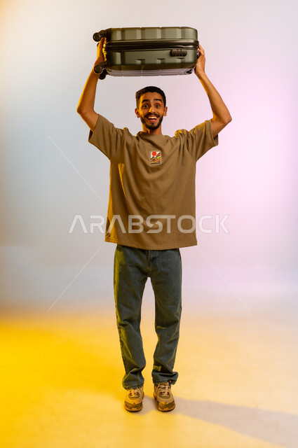 إيماءات وجه تدل على الحماس للقيام برحلة سياحية، تجهيز الأمتعة و الاستعداد للسفر في جولة سياحية, بورتريه لرجل سعودي مبتسم يحمل حقيبة سفر سوداء، شاب عربي اماراتي خليجي يرتدي الزي الكاجوال ينظر الى الكاميرا, الجسم بالطول الكامل، خلفية ملونه