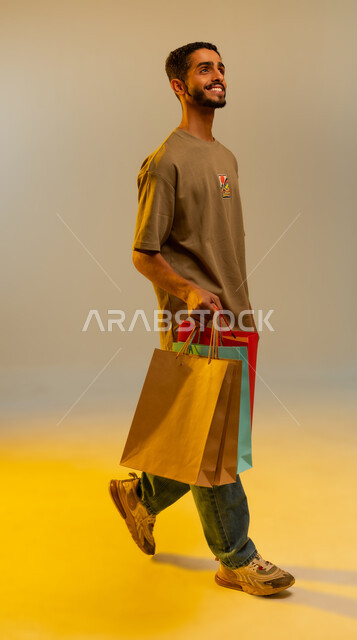 الاستمتاع بالشراء والتبضع،  الاستفادة من العروضات والتخفيضات، بورتريه لرجل عربي خليجي عماني يرتدي الزي الكاجوال يحمل بيده أكياس ورقية, شاب سعودي يقوم بالتبضع وأخذ المستلزمات الضرورية, الجسم بالطول الكامل, خلفية ملونه