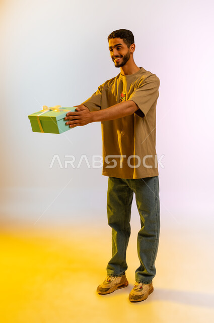 هداءات بمناسبة الأعياد و المناسبات, مناسبات و احتفالات سعيدة, بوتريه لرجل عربي سعودي خليجي يحمل بيده علبة هدايا، إيماءات وجه ويدين تدل على السعاده و السرور، شاب اماراتي يمسك هدية عيد الحب المغلفة، مفاجأة أعياد الميلاد، الجسم بالطول الكامل، خلفية ملونه