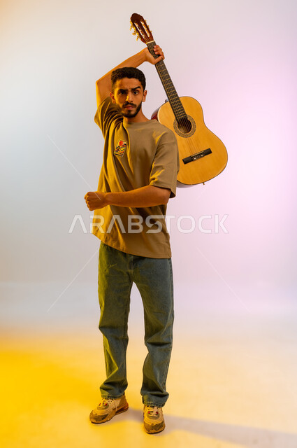 Facial and hand gestures indicating enthusiasm and passion, playing a stringed instrument, portrait of a smiling Saudi Arabian Gulf young man raising the guitar up, Omani young man in casual clothes holding a musical instrument, vocal and artistic musical field, full body, colored background