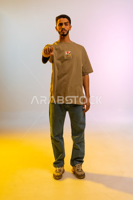 Locating, pointing and explaining something, indicating the location of an item, portrait of a young Arab Gulf Omani man in casual wear, Saudi man pointing his index finger at something he is looking at, full body image, colored background