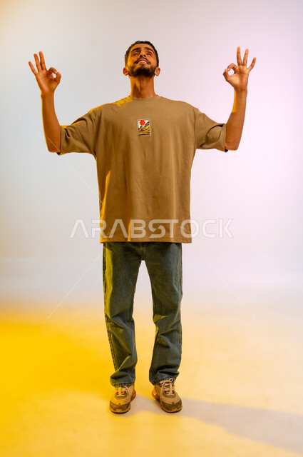 Trying to concentrate and meditate, a Saudi man relaxing and practicing yoga, portrait of a young Arab Gulf Omani man wearing casual clothes raising his hands in gestures of acceptance and approval, expressing quality and perfection, full body, colored background