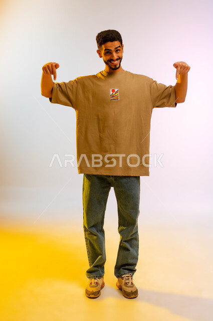 تأكيد حدوث شيء هذه اللحظة, حركات تدل على هذا المكان (هنا)، تحديد موقع معين، بورتريه لشاب عربي خليجي عمانى مبتسم يرتدي الزي الكاجوال ، رجل سعودي  يشير بإصابع السبابة نحو الأسفل, الجسم بالطول الكامل ، خلفية ملونه