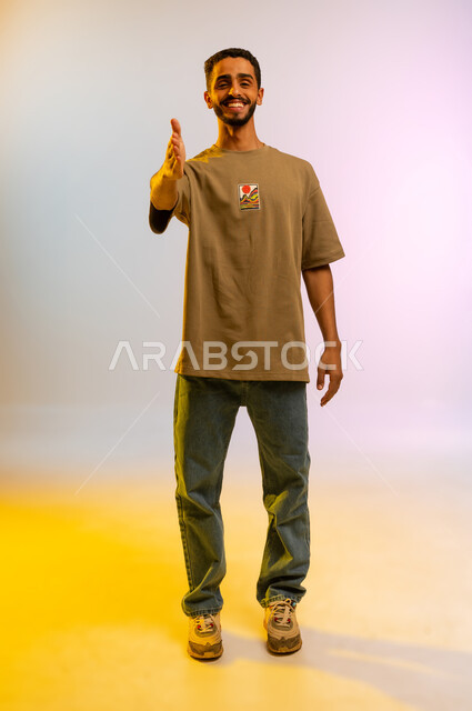 التعارف والصداقة وتكوين العلاقات، مفهوم الاستقبال والترحيب، بورتريه لرجل عربي خليجي عمانى مبتسم يرتدي الزي الكاجوال يمد يده بإيماءات المصافحة، شاب سعودي يقوم بالقاء التحية، الجسم بالطول الكامل، خلفية ملونه
