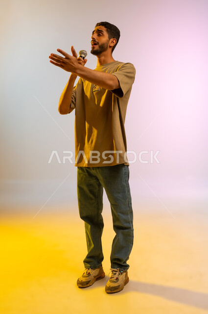 تقديم العروض الصوتية، تعابير الاندماج والتفاعل مع الموسيقا، تسجيل مقطع صوتي غنائي، بورتريه لشاب عربي خليجي عماني مبتسم يرتدي الزي الكاجوال يمسك بيده المايكروفون يقوم بالغناء بإيماءات الفرح والسرور، صورة الجسم بالطول الكامل، خلفية ملونه
