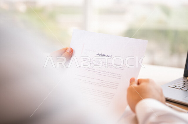 رجل عربي خليجي سعودي يجلس على الحاسوب المحمول ويمسك بيده خطاب توظيف، قراءة خطاب التوظيف، سكرتير يدير الاعمال الورقية، مفهوم التوظيف والعمل، أعمال مكتبية