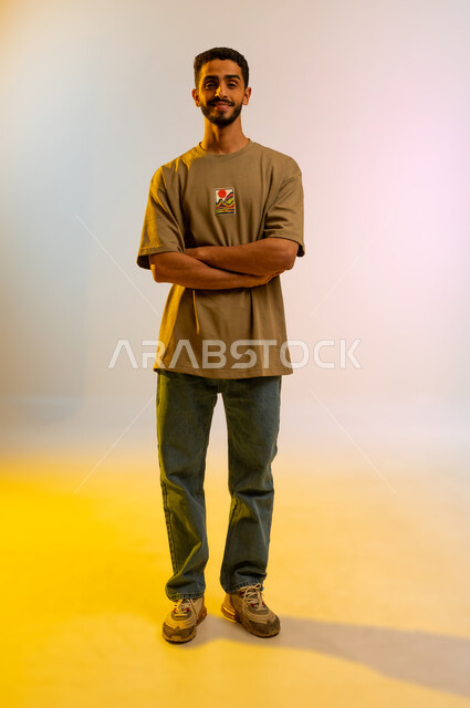 الوقوف باستقامة وثقه بالنفس، بورتريه لشاب وسيم عربي خليجي عماني يرتدي الزي الكاجوال ويقف باستقامة،  شاب سعودي ينظرإلى الكاميرا بإيماءات أيدي متقاطعه، مفهوم الاناقه و الاهتمام بالمظهر الخارجي، صورة الجسم بالطول الكامل، خلفية ملونه