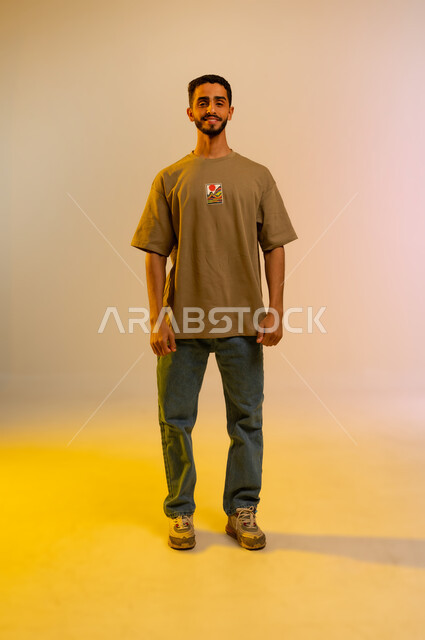 مفهوم الاناقه و الاهتمام بالمظهر الخارجي، الوقوف باستقامة وثقه بالنفس، بورتريه لشاب وسيم عربي خليجي عماني يرتدي الزي الكاجوال ويقف باستقامة،  شاب سعودي ينظرإلى الكاميرا بإيماءات السعادة ، صورة الجسم بالطول الكامل، خلفية ملونه