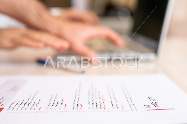 رجل عربي خليجي سعودي يجلس على الحاسوب المحمول وأمامه أوراق خاصة بالسيرة الذاتية، انجاز مهام العمل من خلال الحاسوب المحمول، ادخال بيانات نصية، سكرتير يدير الأعمال الورقية، مفهوم التوظيف والعمل