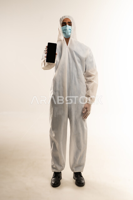 التوعيه من مخاطر كوفيد 19، ارتداء الكمامة لإجراءات الوقاية من فايروس كورونا, بورتريه لطبيب عربي خليجي سعودي يرتدي رداء أبيض بلاستيكي، رجل عربي خليجي عمانى يمسك الهاتف المحمول بشاشة سودء فارغه، الجسم بالطول الكامل، خلفيه بيضاء