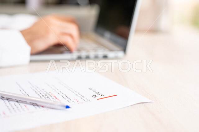 رجل عربي خليجي سعودي يجلس على الحاسوب المحمول وأمامه أوراق خاصة بالسيرة الذاتية، انجاز مهام العمل من خلال الحاسوب المحمول، ادخال بيانات نصية، سكرتير يدير الأعمال الورقية، مفهوم التوظيف والعمل