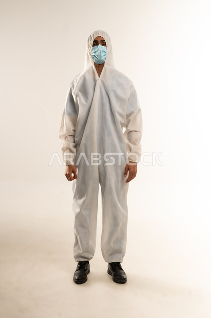 Coronavirus precautions, maintaining safety and avoiding the spread of infection from Covid-19, portrait of a Saudi Arabian Gulf man wearing a white plastic robe and a medical mask looking at the camera, full body image, white background