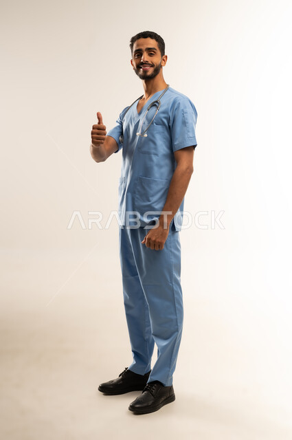 علامة الجودة والتميز، مفهوم الطب والرعاية الصحية، الاستعداد لتقديم الخدمات الطبية للمرضى, ممرض سعودي يعمل بالقطاع الطبي، رفع اصبع الابهام لأعلى بإيماءات القبول, بورتريه لشاب عربي خليجي عماني مبتسم يرتدي يونيفورم العمليقف باستقامة, الجسم بالطول الكامل، خلفية بيضاء