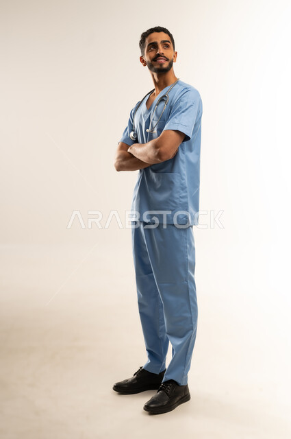 الوقوف بأيدي متقاطعة بتعابير الثقة بالنفس, مفهوم الطب والرعاية الصحية, ممرض سعودي يعمل بالقطاع الطبي، بورتريه لشاب عربي خليجي عماني مبتسم يرتدي يونيفورم العملي يقف باستقامة و ينظر الى شيء ما, الجسم بالطول الكامل، خلفية بيضاء