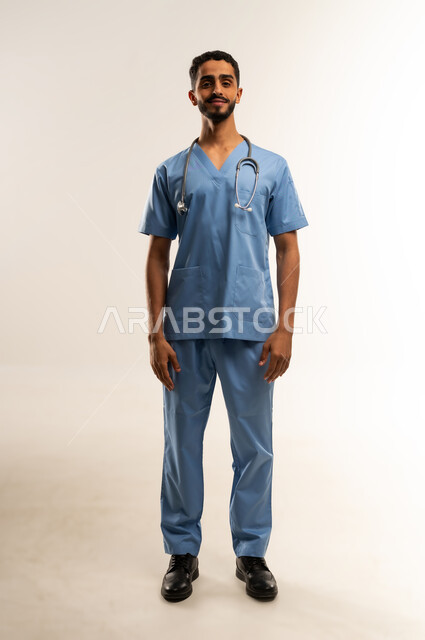 الاستعداد لتقديم الخدمات الطبية للمرضى, مفهوم الطب والرعاية الصحية، ممرض سعودي يعمل بالقطاع الطبي، الوقوف باستعداد والنظر إلى الكاميرا بإيماءات السرور, بورتريه لشاب عربي خليجي عماني مبتسم يرتدي يونيفورم العمل يقف باستقامة, الجسم بالطول الكامل، خلفية بيضاء