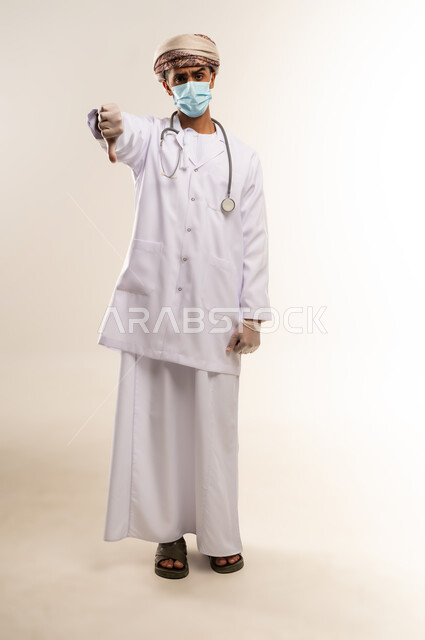 Thumbs down with expressions of anger and dislike, portrait of an Arab Gulf Omani doctor wearing a dishdasha, turban, mask and medical coat, wearing a stethoscope, working in the health sector in the Sultanate of Oman, following safety measures to prevent the Corona virus, full body, white background