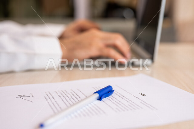 رجل عربي خليجي سعودي يجلس على الحاسوب المحمول وأمامه عقود عمل، انجاز مهام العمل من خلال الحاسوب المحمول، ادخال بيانات نصية، سكرتير يدير الأعمال الورقية، اعمال مكتبية