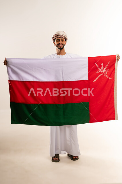 الاعتزاز والفخر بالرموز والراية العمانية، الاحتفال بذكرى اليوم الوطني العماني، المشاركة بالاعياد والمناسبات الوطنية، بورتريه لشاب عربي خليجي عماني يرتدي الدشداشة والعمامة ، مفهوم حب الوطن والإنتماء إليه، الجسم بالطول الكامل، خلفية بيضاء