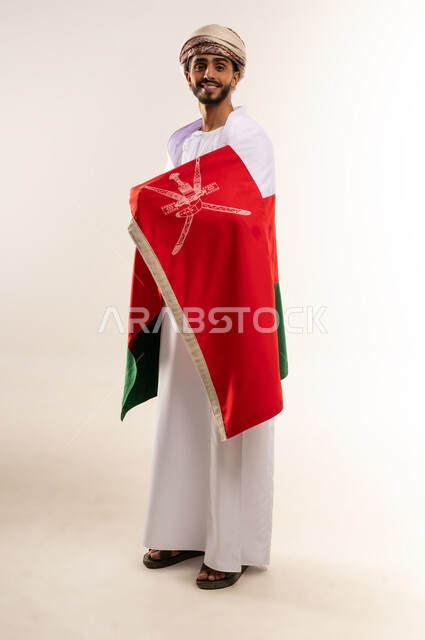 الاحتفال بذكرى اليوم الوطني العماني، المشاركة بالاعياد والمناسبات الوطنية، الاعتزاز والفخر بالرموز والراية العمانية، بورتريه لشاب عربي خليجي عماني يرتدي الدشداشة والعمامة ، مفهوم حب الوطن والإنتماء إليه، الجسم بالطول الكامل، خلفية بيضاء