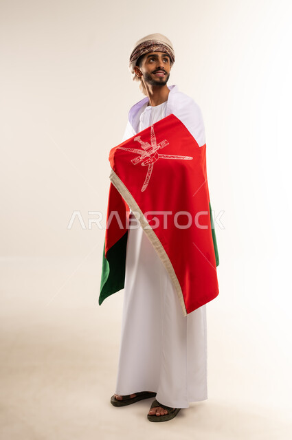 Participating in national holidays and occasions, celebrating Omani National Day, and taking pride in Omani symbols and the Omani flag. Portrait of a young Gulf Arab Omani man wearing a dishdasha and turban. The concept of patriotism and belonging to the homeland. Full-length body, white background.