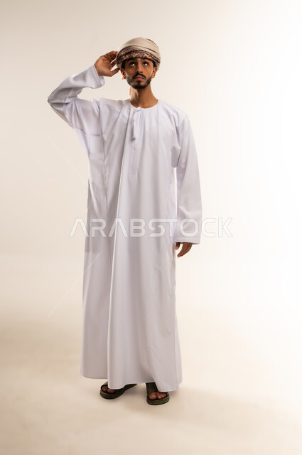 Trying to see distant objects, gestures of concentration for clear vision, the concept of looking to the future, a portrait of a young Arab man from the Gulf, Omani, wearing a dishdasha and turban, placing his hand on his forehead and looking in a certain direction, full-length body, white background