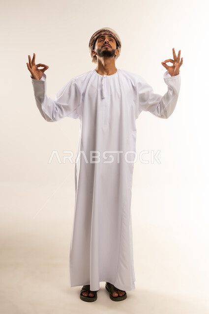 علامة الجوده و الإمتياز والتميز, مفهوم القبول والموافقه، بورتريه لشاب عربي خليجي عماني مبتسم يرتدي الدشداشة والعمامة ينظر الى الاعلى يرفع يديه للاعلى بإيماءات الإعجاب والرضا، صورة الجسم بالطول الكامل, خلفية بيضاء
