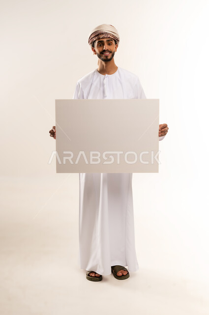 ايماءات السعادة والسرور، إعلانات وعروض تسويقية، استخدام لوحات توضيحية ووسائل تعليمية، بورتريه لرجل عربي خليجي عماني مبتسم يرتدي الدشداشه والعمامة يحمل بيده لوح ابيض كبير (موك اب فارغ)، الطول الكامل للجسم، خلفية بيضاء.