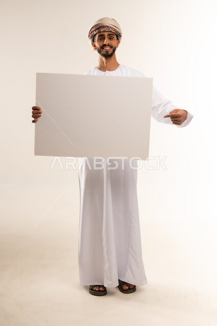 إعلانات وعروض تسويقية، استخدام لوحات توضيحية ووسائل تعليمية، بورتريه لرجل عربي خليجي عماني مبتسم يرتدي الدشداشه والعمامة يحمل بيده لوح ابيض كبير (موك اب فارغ)، ايماءات السعادة والسرور، الطول الكامل للجسم، خلفية بيضاء.