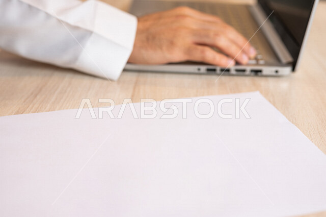 رجل عربي خليجي سعودي يجلس على الحاسوب المحمول وامامه مجموعة من الأوراق الفارغة، ادارة الاعمال من خلال الحاسوب المحمول، ادخال البيانات من خلال لوحة المفاتيح،  محتويات نصية