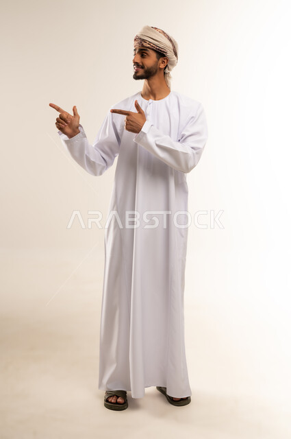 Looking aside with gestures of pleasure and pointing at something, portrait of an old Arab Gulf Omani man wearing a dishdasha and traditional Omani turban standing straight, concern for outward appearance, full body, white background