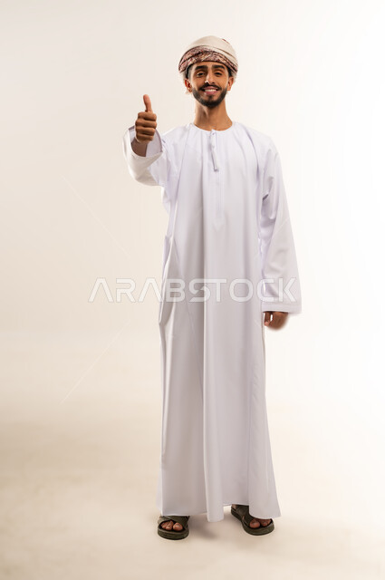 تعابير الموافقة والرضى والقبول، بورتريه لشاب عربي خليجي عماني مبتسم يرتدي الدشداشة و العمامة يقف و يرفع اصبع الابهام للاعلى بإيماءات الاعجاب والسرور، صورة الجسم بالطول الكامل، خلفية بيضاء